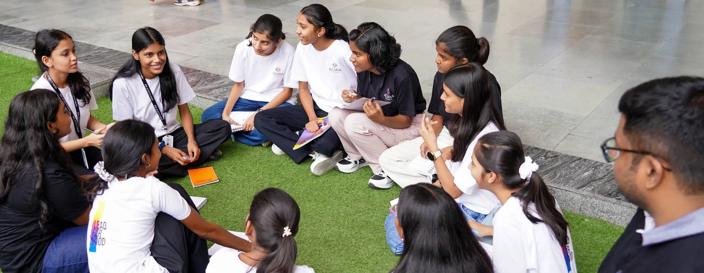 Students sitting in a circle on the grass, talking, with notebooks in their laps